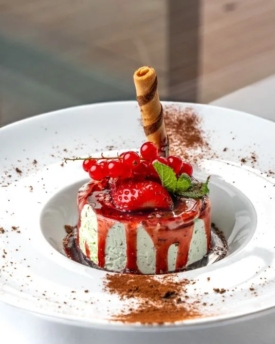 Dolce in un piatto bianco con ribes, fragola, menta e una cialda arrotolata, cacao spolverato sul bordo.