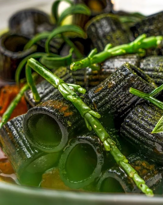 Paccheri scuri in primo piano con verdure verdi sottili.