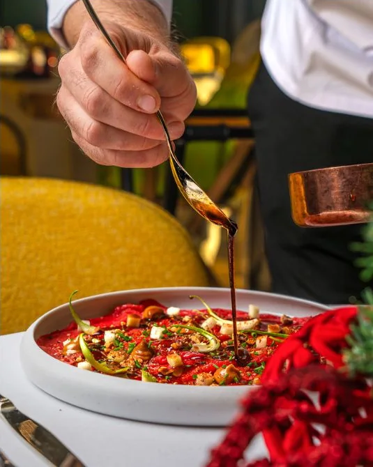 Un cameriere versa una salsa scura con un cucchiaio su un piatto rosso, al tavolo.